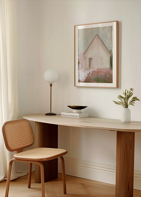 ein moderner, minimalistischer Arbeitsplatz mit einem Holzschreibtisch, einem geflochtenen Stuhl, einer Lampe und einem gerahmten Gemälde eines Hauses.